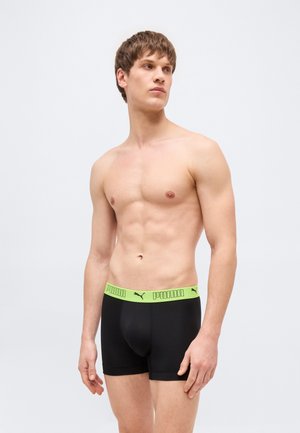 Jeune homme torse nu portant un boxer Puma noir avec une ceinture verte, debout devant un fond clair uni, regardant vers la droite.