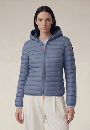 Kvinde iført en blå quiltet hættetrøjejakke med orange lynlåstræk og et logopatch på ærmet, kombineret med hvide bukser.