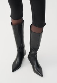 Black leather knee-high boots with pointed toes and zipper details, featuring a sleek design and glossy texture.