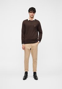 Pull marron avec un col rond et un petit logo doré, porté par-dessus une chemise blanche à col, associé à un pantalon beige retroussé et des bottes noires.