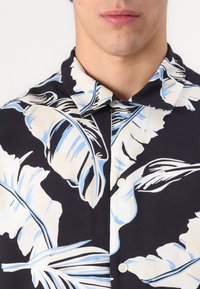 Camicetta a maniche corte in tessuto nero con grandi motivi di foglie beige e azzurro chiaro; presenta un colletto standard e chiusura frontale con bottoni.