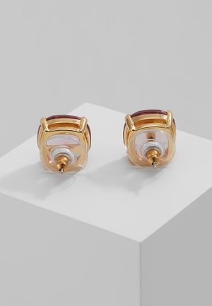 Boucles d'oreilles clous en or avec pierres précieuses rouges carrées et attaches en plastique transparent exposées sur un socle blanc sur fond gris.
