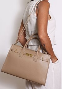 Femme en haut sans manches blanc portant un grand sac en cuir beige avec une fermeture dorée et une sangle ajustable sur l'épaule.