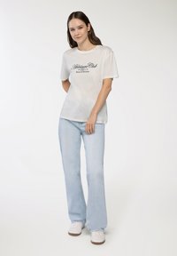 Biała bawełniana koszulka z napisem "Athleisure Club" oraz "Relax & Wellness" w kolorze czarnym. Noszona z jasnoniebieskimi dżinsami typu flare i białymi trampkami.