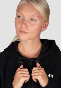 Melns kapuces džemperis ar rāvējslēdzēja aizdari; tam ir gluda virsma un logo uz krūtīm. Modelis tur rāvējslēdzēju ar abām rokām.