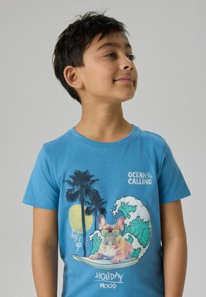 Boy with short dark hair wears blue t-shirt featuring a dog with sunglasses on a surfboard, palm trees, waves, sun, and text "Ocean Calling."