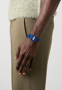 Montre numérique bleue avec un cadran rectangulaire, bracelet en caoutchouc et affichage numérique montrant l'heure. Portée sur une main à la peau brune, assortie à un pantalon beige.