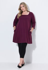 Bordeauxfarvet tunika kjole med trekvartærmer, lige snit, lommer, parret med sorte leggings og sorte flade sko, positioneret mod en hvid baggrund.