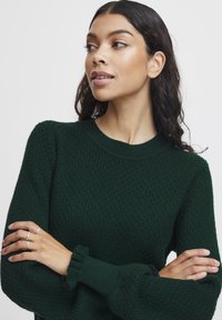 Dark green knitted sweater with a textured pattern, ribbed crew neckline, and ruffled cuffs. Model has her arms crossed and is facing left.