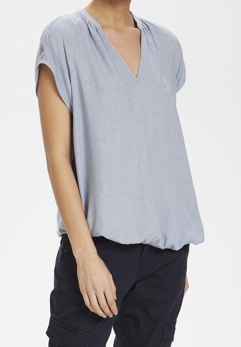 Ljusblå kortärmad blus med V-ringning, rynkade axeldetaljer och en lös passform som faller något vid fållen.