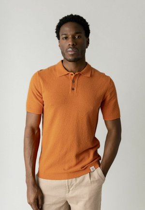 Mees lühikeste lokkis juustega, kes kannab oranži lühikeste varrukatega tekstuuriga polosärki ja heledaid beeže pükse, seisab ühe käega pihus.