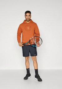 Bränd orange hoodie med logotyp i mitten, svarta sportshorts, svarta sneakers och håller i ett tennisracket med blå accenter.