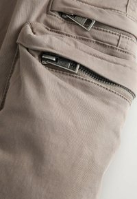 Un tissu beige clair avec une texture lisse présente des poches zippées angulaires. Le matériel en métal argenté du zip ajoute du détail au design.