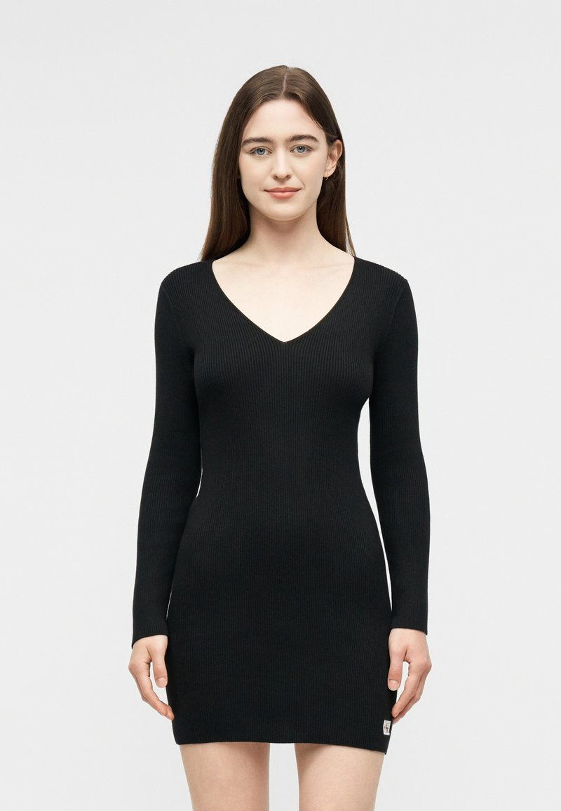 Νεαρή γυναίκα με μακριά καστανά μαλλιά, φορώντας εφαρμοστό μακρυμάνικο μαύρο ριμπ φόρεμα με λαιμόκοψη σε σχήμα V, στέκεται μπροστά σε απλό φωτεινό φόντο.