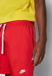 Personne à la peau foncée portant un short Nike rouge vif avec un cordon blanc et un t-shirt jaune, debout devant un fond uni.