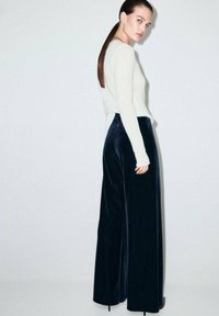 Pantaloni largi din catifea bleumarin asociați cu un pulover ajustat de culoare deschisă. Textură netedă, design lejer și siluetă lungă evidențiate.