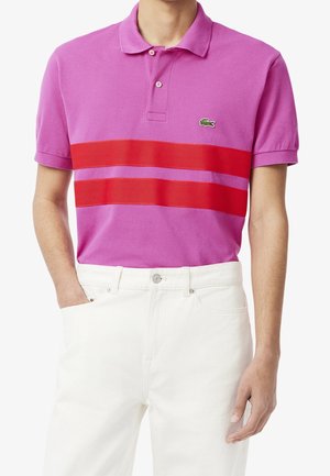 Homme portant un polo rose avec deux bandes rouges horizontales et un pantalon blanc, main dans la poche sur fond blanc.