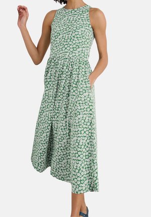 Robe midi verte sans manches avec imprimé marguerites blanches, taille cintrée, encolure ronde, poches latérales, portée avec des chaussures plates bleues.