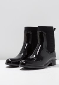 Black ankle rain boots made of shiny rubber. Features elastic side panels, a loop at the back, and a low, textured heel. Smooth finish.