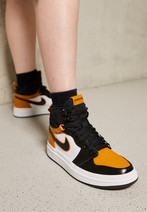 Osoba nosząca czarno-biało-pomarańczowe wysokie sneakersy Nike Air Jordan z czarnymi sznurówkami i czarnymi skarpetkami na jasnej, cętkowanej podłodze.