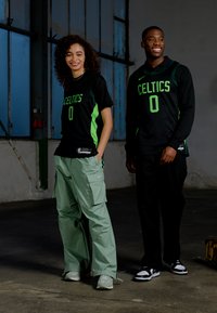 Svart basketballdrakter har neongrønn "CELTICS" skrift og nummer "0." Kvinnen har på seg lysegrønne cargo-bukser; mannen har på seg svarte bukser.