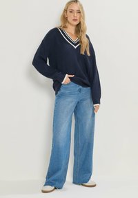 Granatowy sweter dzianinowy z dekoltem w serek i białymi paskami; połączony z jasnoniebieskimi spodniami typu wide-leg; modelka stoi z rękami w kieszeniach.