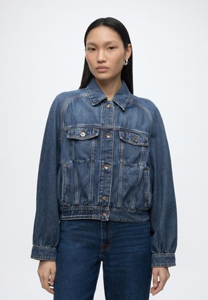 Vrouw met een blauwe spijkerjas met knopen en donkerblauwe jeans, staand tegen een effen lichte achtergrond.