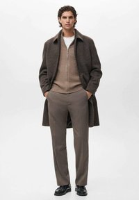 Uomo che indossa un lungo cappotto a quadri marrone sopra un maglione beige con zip, pantaloni taupe e scarpe di pelle nere, in piedi con le mani nelle tasche.