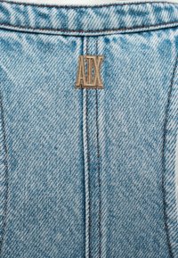 Υφασμάτινο τζιν ανοιχτού μπλε χρώματος με κάθετες ραφές και ένα μικρό καφέ κεντητό έμβλημα με τα γράμματα "ADX" κοντά στο πάνω κέντρο.