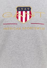 Sudadera gris con un emblema de escudo con rayas rojas y blancas, letras doradas "GANT 1949" y "AMERICAN SPORTSWEAR". Textura suave.