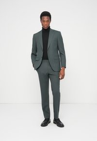 Σκούρο πράσινο κοστούμι με στενή γραμμή, σακάκι με δύο κουμπιά, πέτα με εγκοπές και παντελόνι με ίσια μπάση, σε συνδυασμό με μαύρο ζιβάγκο και μαύρα παπούτσια.