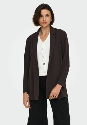 Femme aux cheveux courts et bruns portant un blazer marron foncé sur un haut blanc boutonné et un pantalon noir sur un fond clair uni.
