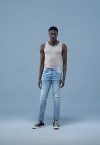 Világosszürke trikó, világoskék farmernadrág és fekete magas szárú tornacipők. Álló póz világoskék háttér előtt.