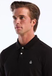 Zwarte poloshirt met een gestructureerde afwerking, drie-knopenlijst en een klein wit pinguïnlogo op de linkerkant van de borst.