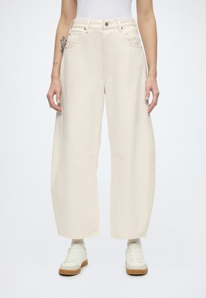 Persona che indossa pantaloni crema a vita alta e gamba larga con tasche frontali, calzini bianchi, scarpe da ginnastica bianche e una canotta bianca senza maniche.