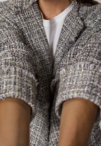 Wyróżniająca się teksturowana, tkana marynarka w kolorach niebieskim, kremowym i beżowym. Posiada postrzępione krawędzie oraz luźny krój, noszona na białym t-shircie.