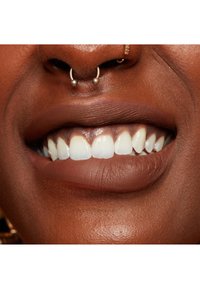 Närbild av en mun med fylliga läppar i en djup brun nyans, med vita tänder och en septum-piercing med två metallkulor.