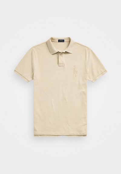 Beige színű pólóing, pamutból készült, háromgombos gallérral, rövid ujjakkal és hímzett logóval a bal mellrészen. Sima tapintású.