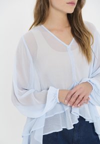 Šviesiai mėlyna, permatoma bluzelė su V formos iškirpte, sluoksniuotomis raukinėmis ir elastingais rankogaliais. Audinys turi subtilią tekstūrą, kuri sukuria plazdantį siluetą.