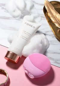 Nettoyant micro-mousse FOREO dans un tube blanc avec des accents métalliques, aux côtés d'un dispositif de nettoyage en silicone rose, entouré de bulles de savon.