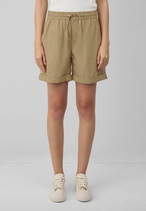 Shorts beiges arrivés aux genoux avec des revers, taille élastique et poches avant. Associés à des baskets blanches sur un fond neutre.