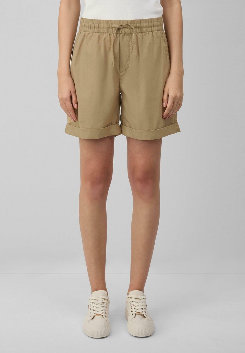 Beige, knielange shorts met opgerolde mouwen, elastische tailleband en voorzakken. Gecombineerd met witte sneakers op een neutrale achtergrond.