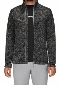 Uomo che indossa una giacca con zip nera e grigia a textures sopra una camicia nera stampata e pantaloni grigio chiaro, in piedi con le braccia rilassate.