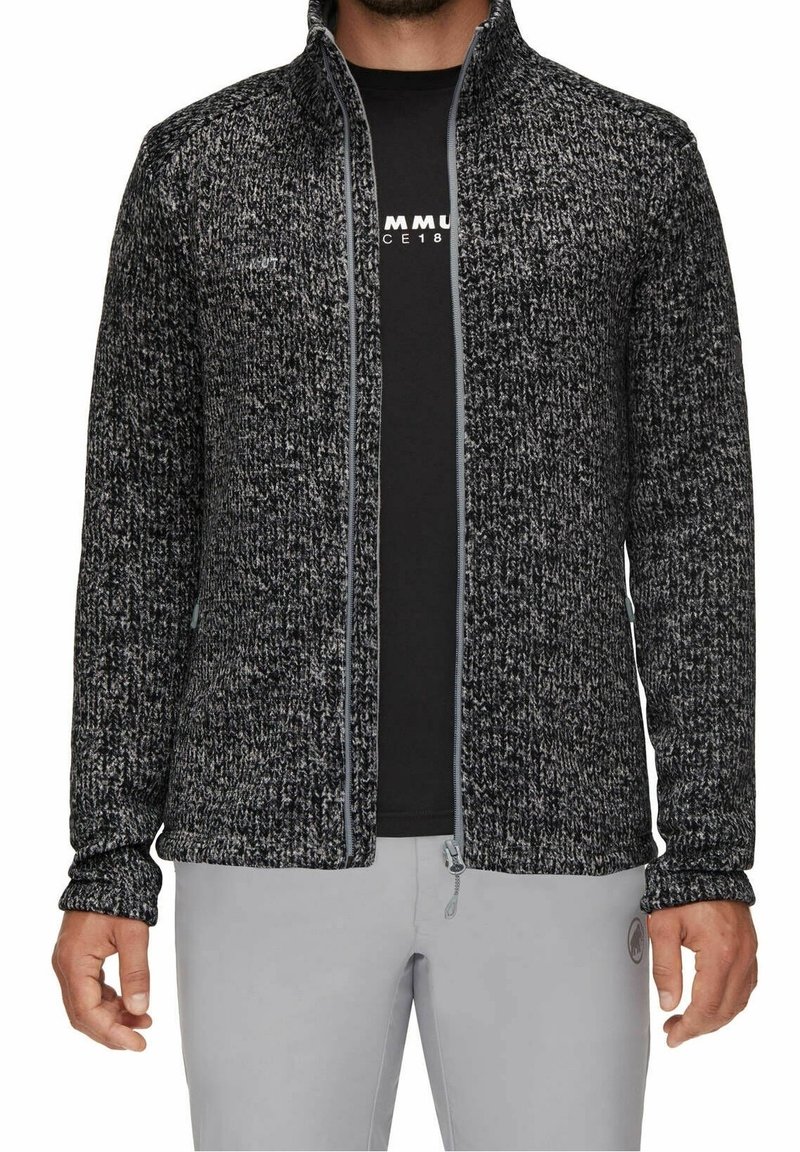 Uomo che indossa una giacca con zip nera e grigia a textures sopra una camicia nera stampata e pantaloni grigio chiaro, in piedi con le braccia rilassate.