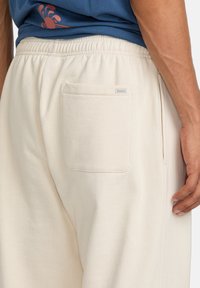 Helle beigefarbene Sweatpants mit einem elastischen Bund, Seitentasche und einer Gesäßtasche mit einem dezenten Markenlabel. Glatte Stoffoberfläche.