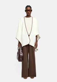 Biała wełniana ponczo z brązowymi lamówkami, połączona z brązowymi spodniami szerokimi i ciemnobrązową torbą z srebrnym okuciem. Model nosi oversize'owe okulary.