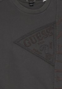 Felpa grigia scura con un patch triangolare testurizzato che presenta il logo "GUESS", cuciture intricate e un leggero effetto invecchiato.
