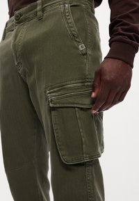 Cargo bukser i olivengrøn med ternet struktur, frontlommer og sidelomme til cargo med klapdetalje. Synligt metalhardware.
