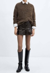 Maglione marrone fluffoso, camicia bianca con colletto sotto, gonna nera plissettata in ecopelle, collant beige, stivali neri in pelle al ginocchio.