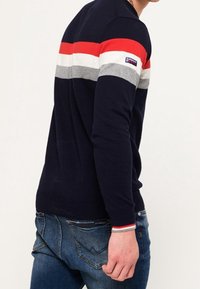 Tmavě modrý svetr s červenými, bílými a šedými vodorovnými pruhy přes ramena. Žebrovaný lem a manžety. Kombinován s džínami modré barvy.